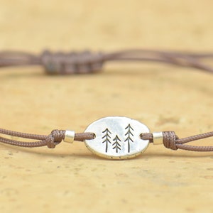 Puede incluir: Pulsera de cordón marrón con un colgante ovalado plateado con tres árboles grabados. La pulsera tiene nudos corredizos ajustables y pequeñas cuentas plateadas. El diseño es sencillo e inspirado en la naturaleza.