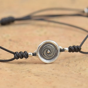 Può includere: Un braccialetto in corda nera con un ciondolo circolare argentato che presenta un design floreale stilizzato.