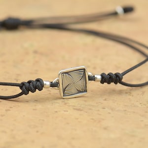 Può includere: Un braccialetto regolabile in corda nera con un ciondolo quadrato argentato con un motivo geometrico.