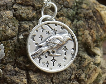 May include: A silver pendant necklace featuring a detailed raven design within a circular frame. The raven is surrounded by stars and a crescent moon. The pendant hangs from a delicate silver chain, set against a textured, brown background.