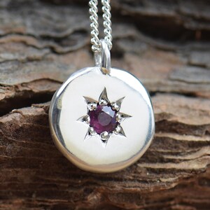 Könnte beinhalten: Eine silberne Halskette mit einem runden Anhänger, der ein Sternenmuster mit einem kleinen violetten Edelstein in der Mitte zeigt.