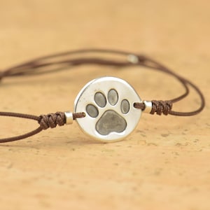 May include: A bracelet featuring a circular silver-toned charm with an embossed paw print design. The charm is centered on a thin, dark brown cord with adjustable sliding knots and small silver-toned metal beads on either side of the charm.