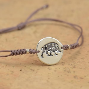Puede incluir: Un brazalete de cordón marrón con un dije de plata que presenta un diseño detallado de jabalí.
