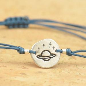Könnte beinhalten: Ein blaues Armband aus Schnur mit einem silbernen Anhänger, der einen Planeten mit Ringen und Sternen zeigt.