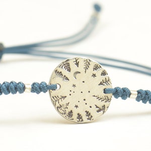 May include: A blue cord bracelet with a silver charm featuring a moon, stars, and trees.