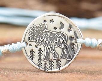 May include: A silver bracelet with a round charm featuring a moose silhouette, stars, and a crescent moon. The charm is surrounded by a light blue cord.
