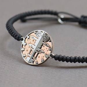 May include: A black cord bracelet with a silver and rose gold circular charm. The charm has a detailed design of a city skyline and stars.