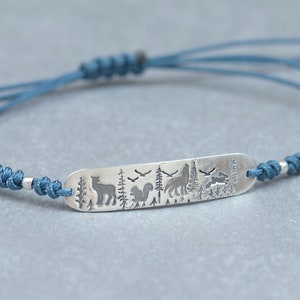 Pode incluir: Um bracelete de cordão azul com um amuleto de prata com um veado, um esquilo e árvores.