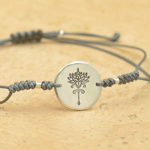 May include: A gray adjustable cord bracelet with a silver circular charm featuring an engraved lotus flower design.
