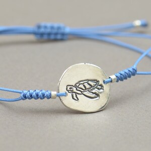Könnte beinhalten: Ein verstellbares Armband aus blauem Kordel mit einem silbernen Anhänger in Form einer Schildkröte.