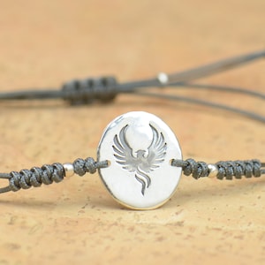 May include: A grey cord bracelet with a silver charm featuring a phoenix design.