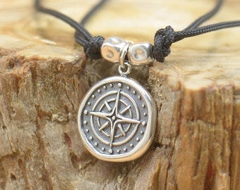 Può includere: Una collana con un ciondolo a forma di bussola d'argento su un cordoncino nero. La bussola è rotonda e presenta un design dettagliato con una stella al centro.
