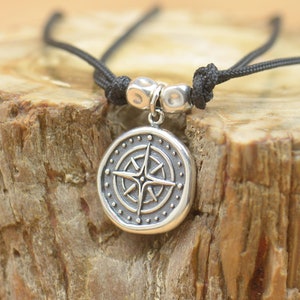 Può includere: Una collana con un ciondolo a forma di bussola d'argento su un cordoncino nero. La bussola è rotonda e presenta un design dettagliato con una stella al centro.