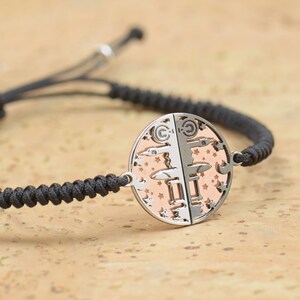 May include: A black braided cord bracelet with a silver and rose gold circular pendant featuring a detailed design of stars and geometric shapes.
