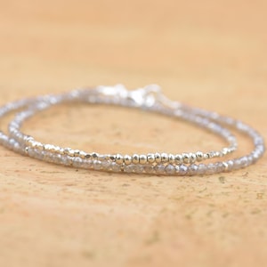 May include: A delicate silver bracelet with two strands of small, shimmering beads. One strand features a series of tiny silver beads, while the other is composed of faceted, light grey beads. The bracelet is designed to wrap around the wrist.