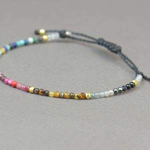 Könnte beinhalten: Ein farbenfrohes Perlenarmband mit schwarzem Band und goldenen Akzenten. Das Armband ist aus verschiedenen Edelsteinen gefertigt, darunter Türkis, Amethyst und Rubin.