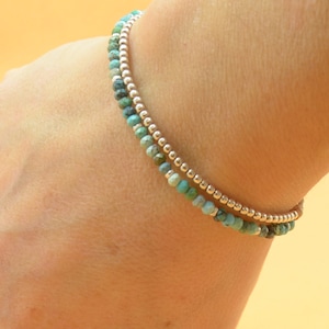May include: Two bracelets stacked on a wrist. The top bracelet is made of silver beads. The bottom bracelet is made of turquoise beads.