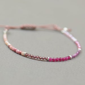 Könnte beinhalten: Ein rosafarbenes und rotes Edelstein-Armband mit einem silbernen Verschluss und einer rosafarbenen Schnur. Das Armband besteht aus kleinen runden Perlen und hat einen Farbverlauf.