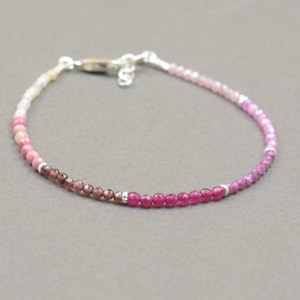 May include: A delicate bracelet with a gradient of pink, red, and brown gemstones. The bracelet is made with small, round beads and has a silver clasp.
