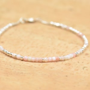 Puede incluir: Un delicado brazalete hecho con pequeñas cuentas blancas y rosadas y detalles plateados.