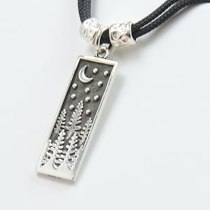 Puede incluir: Un collar con un colgante de plata rectangular que presenta una luna creciente, estrellas y una escena de bosque estilizada.