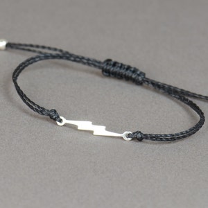 Op de afbeelding: Een armband van zwart koord met een zilveren bliksem-bedel.