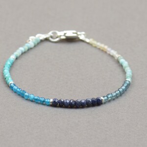 Puede incluir: Un delicado brazalete con un degradado de cuentas de piedras preciosas azules. El brazalete tiene un cierre plateado y una mezcla de cuentas azul claro, turquesa y azul oscuro.