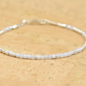 Puede incluir: Un delicado brazalete hecho con pequeñas cuentas de piedra lunar blanca y cuentas de plata. El brazalete está enhebrado en una cadena de plata.