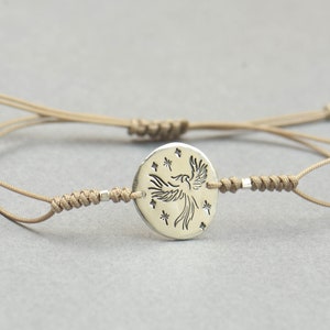 Pu&ograve; includere: Un braccialetto in corda beige con un ciondolo d'argento che presenta una fenice stilizzata con stelle intorno.