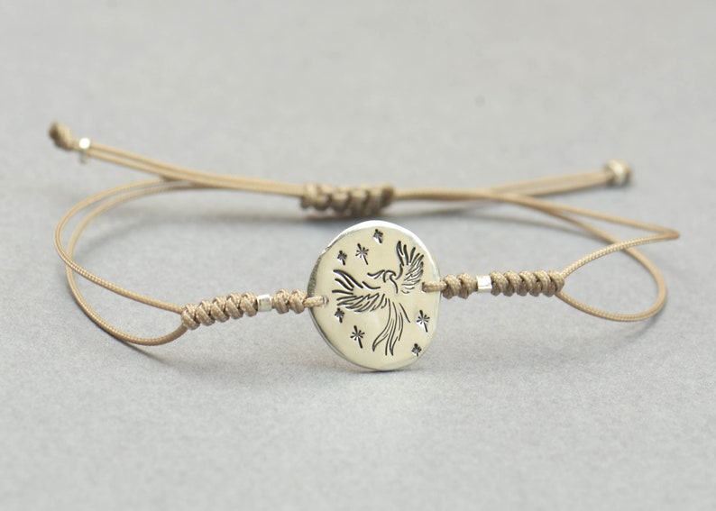 Pu&ograve; includere: Un braccialetto in corda beige con un ciondolo d'argento che presenta una fenice stilizzata con stelle intorno.