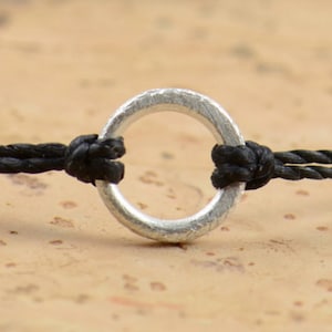 May include: A black cord bracelet with a silver ring charm. The ring is textured and has a smooth, rounded finish.