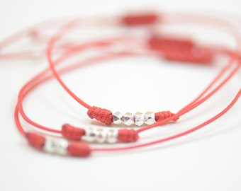 May include: Three red string bracelets with silver beads. The bracelets are tied in a knot and are lying on a white surface.