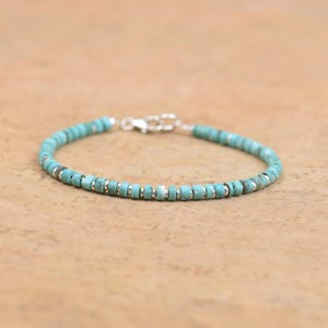 May include: A turquoise beaded bracelet featuring small, round beads and silver-coloured spacers. The bracelet is secured with a silver-coloured clasp. The beads are approximately 0.5 cm in diameter. The bracelet is displayed on a light brown surface.