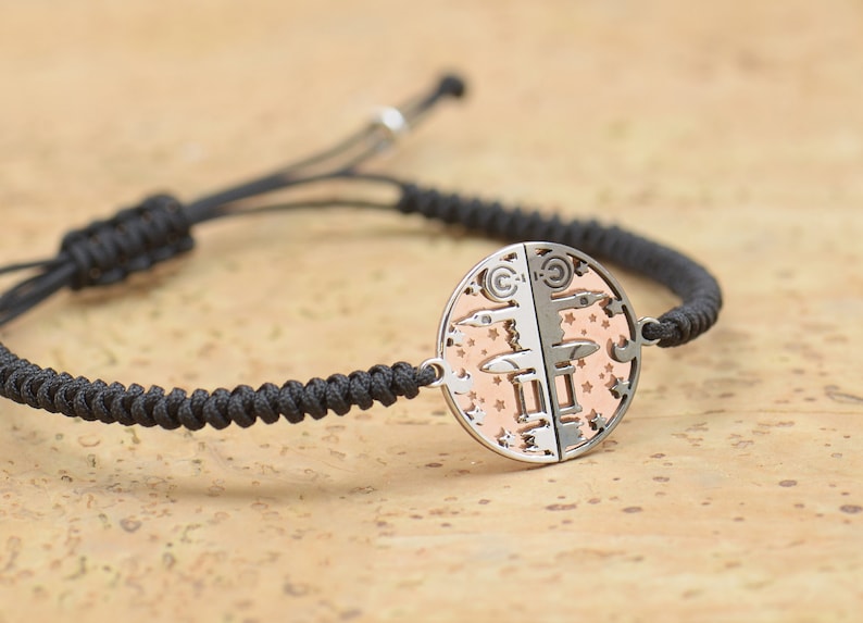May include: A black braided cord bracelet with a silver and rose gold circular pendant featuring a detailed design of stars and geometric shapes.