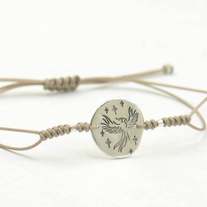 Pu&ograve; includere: Un braccialetto in corda beige con un ciondolo circolare argentato che presenta un design stilizzato di fenice con stelle.