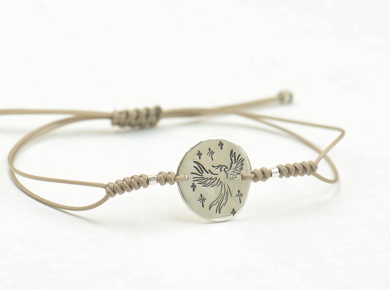Pu&ograve; includere: Un braccialetto in corda beige con un ciondolo circolare argentato che presenta un design stilizzato di fenice con stelle.