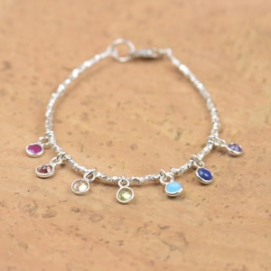 Puede incluir: Una pulsera de plata con una cadena texturizada y pequeños dijes de piedras preciosas de colores. Los dijes incluyen piedras rosas, moradas, verdes, azules y transparentes. La pulsera se exhibe sobre una superficie marrón claro.