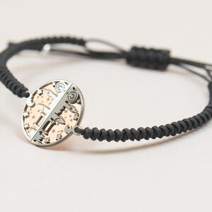 May include: A black braided cord bracelet with a silver and rose gold circular charm featuring a zodiac symbol and stars.