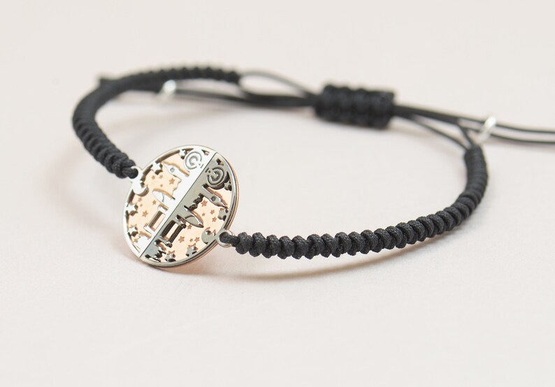 May include: A black braided cord bracelet with a silver and rose gold circular charm featuring a zodiac symbol and stars.