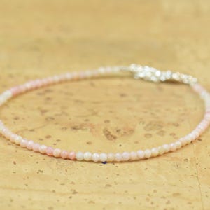 May include: A delicate bracelet made of small, round, pink and white opal beads. The bracelet is strung on a silver chain.