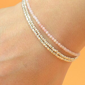 May include: A layered bracelet with three strands of beads. The top strand is made of small, pale pink beads. The bottom two strands are made of silver beads.