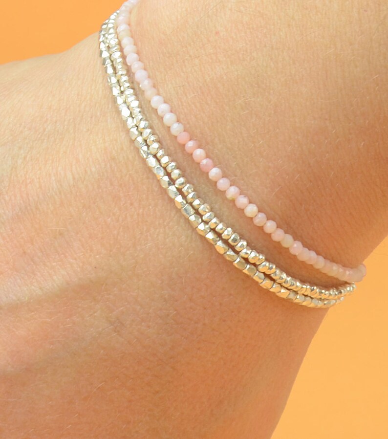 May include: A layered bracelet with three strands of beads. The top strand is made of small, pale pink beads. The bottom two strands are made of silver beads.