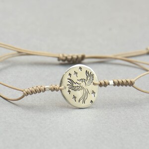 Pu&ograve; includere: Un braccialetto in corda beige con un ciondolo d'argento che presenta una fenice stilizzata con stelle intorno.