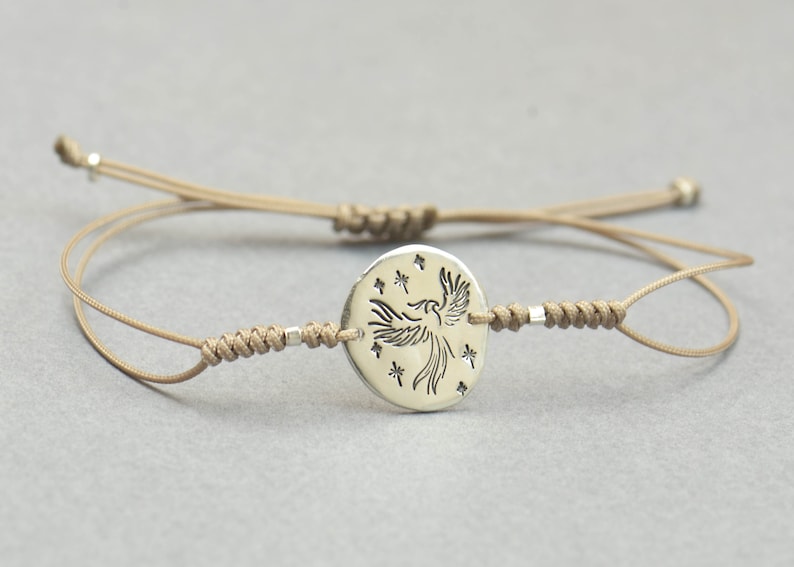 Pu&ograve; includere: Un braccialetto in corda beige con un ciondolo d'argento che presenta una fenice stilizzata con stelle intorno.