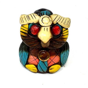 May include: A colorful owl figurine with red gem eyes, a cream-colored beak, and yellow, blue, and pink accents. The owl has a dark brown body and is likely made of carved wood or a similar material. The figurine is a decorative item.