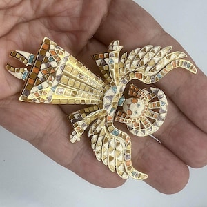 May include: A gold-toned brooch with a mosaic design featuring a fan-like shape and stylized wings. The mosaic incorporates squares and triangles in shades of beige, orange, brown, and blue. The brooch is held in a hand.