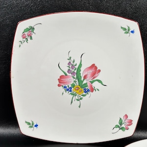 May include: A white square plate with a red rim features a hand-painted floral design. The design includes pink tulips, blue and yellow flowers, and green leaves.