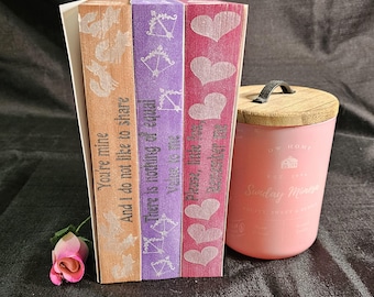 May include: A collection of decorative wooden bookends in shades of peach, purple, and pink, each with a different romantic quote. A pink candle with a wooden lid and the text "Sunday Mine" is next to the bookends.