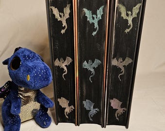 May include: Three black books with a dragon silhouette design on each cover. The dragons are in different colors, including blue, green, and gold. The books are stacked on top of each other.