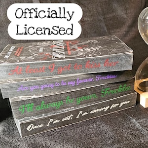 May include: A stack of three books with black covers and colorful text. The top book reads "At least I got to kiss her" in red. The middle book reads "Are you going to be my forever, Freckles" in purple. The bottom book reads "I'll always be yours, Freckles" in green.